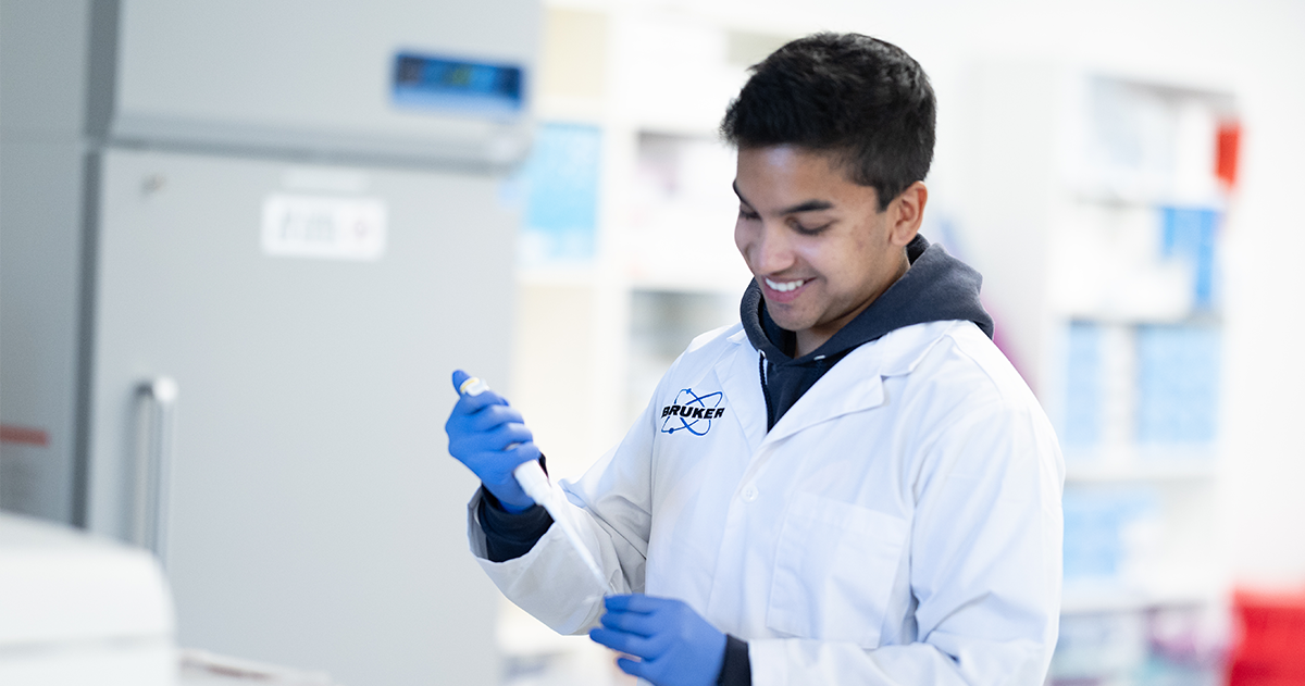 A smiling scientist wearing a Bruker lab coat holds a pipette in a lab